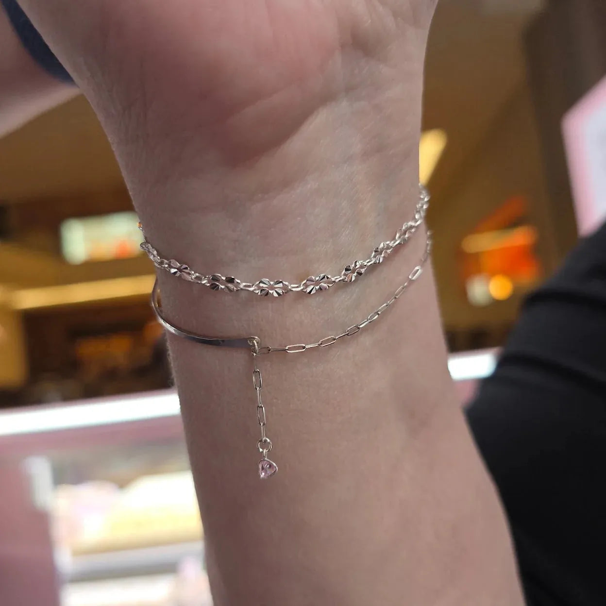 A close-up of a person's wrist wearing two delicate silver bracelets, including a floral chain bracelet and a thin bangle with a dangling charm, set against a blurred indoor background.