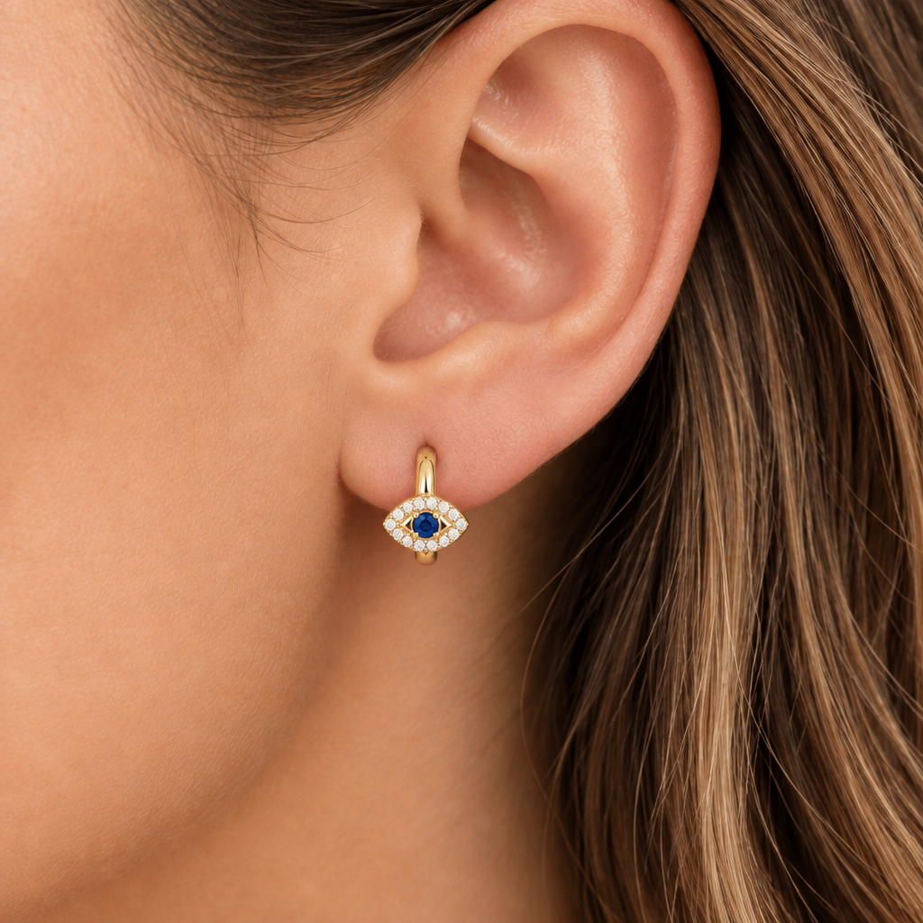 A close-up of a model's ear wearing a small gold huggie hoop earring with an evil eye motif. The earring features a blue oval stone in the center, surrounded by small clear stones, and is shown against the model's skin and brown hair.