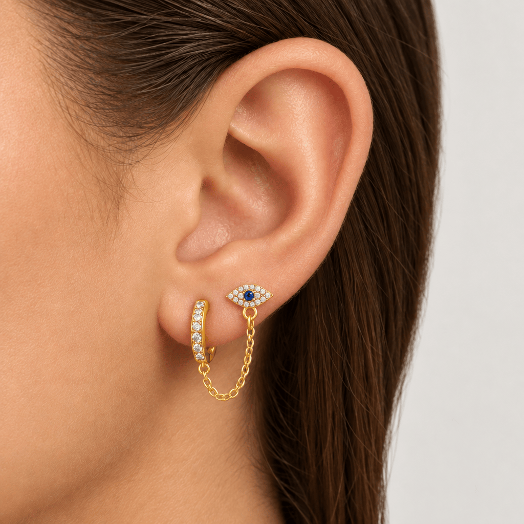 A close-up of a person's ear wearing a gold huggie hoop earring with a dangling chain and an eye-shaped stud earring set with clear stones and a blue center, against a light gray background.
