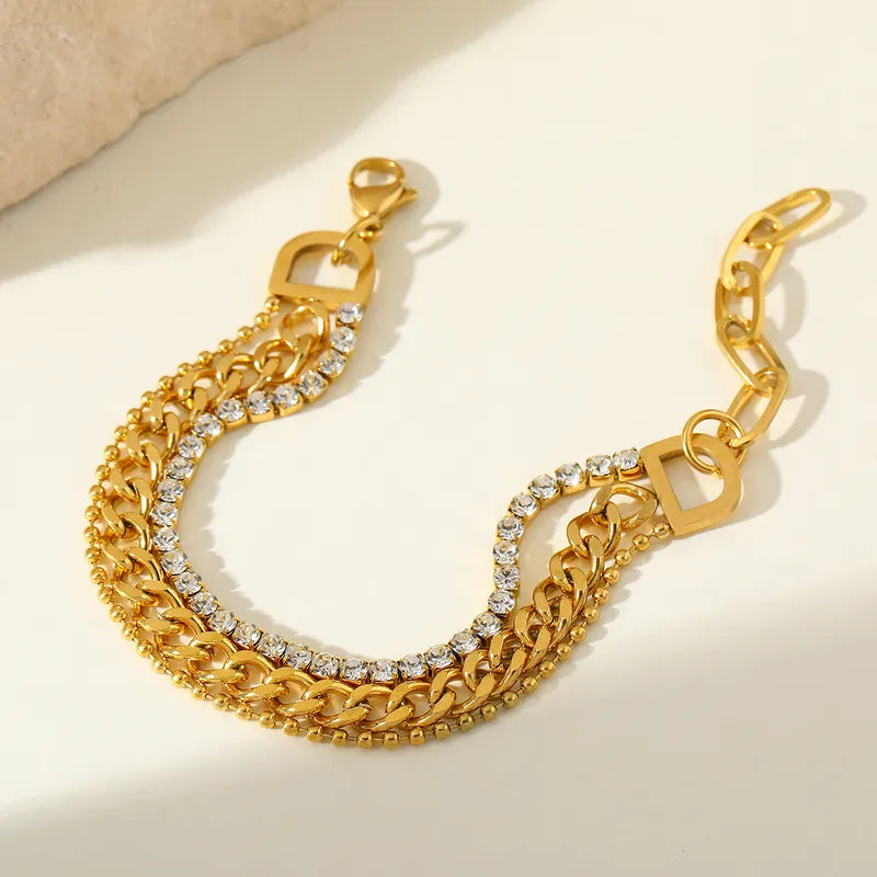 A gold-toned chain bracelet featuring multiple layered curb links, with one row set with clear rhinestones, displayed on a light cream surface.