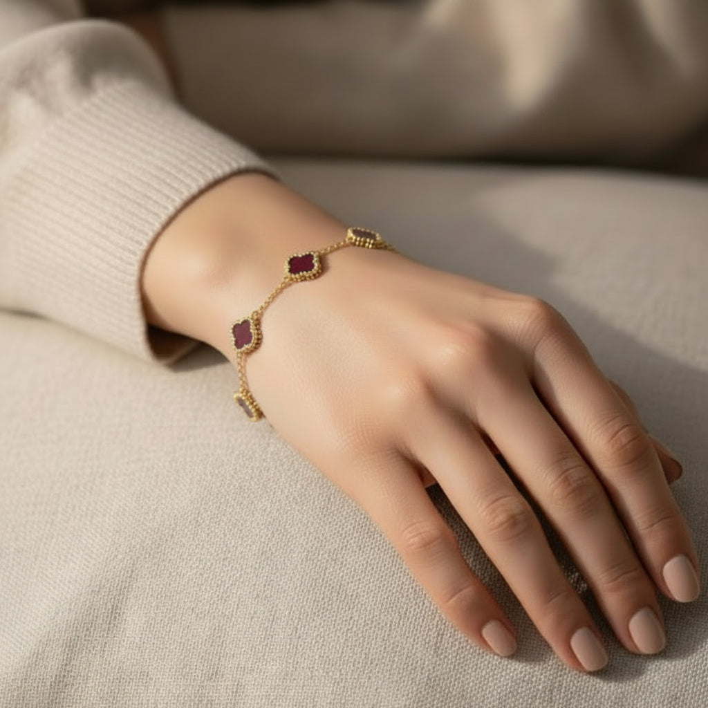 A delicate gold chain bracelet with three small rectangular purple stones, worn on a woman's wrist. The person is wearing a cream-colored ribbed knit sweater, and the background is a soft, textured neutral fabric.
