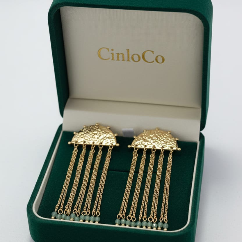 A pair of gold-toned dangling earrings displayed in an open dark green jewelry box with the brand name CinloCo printed inside.