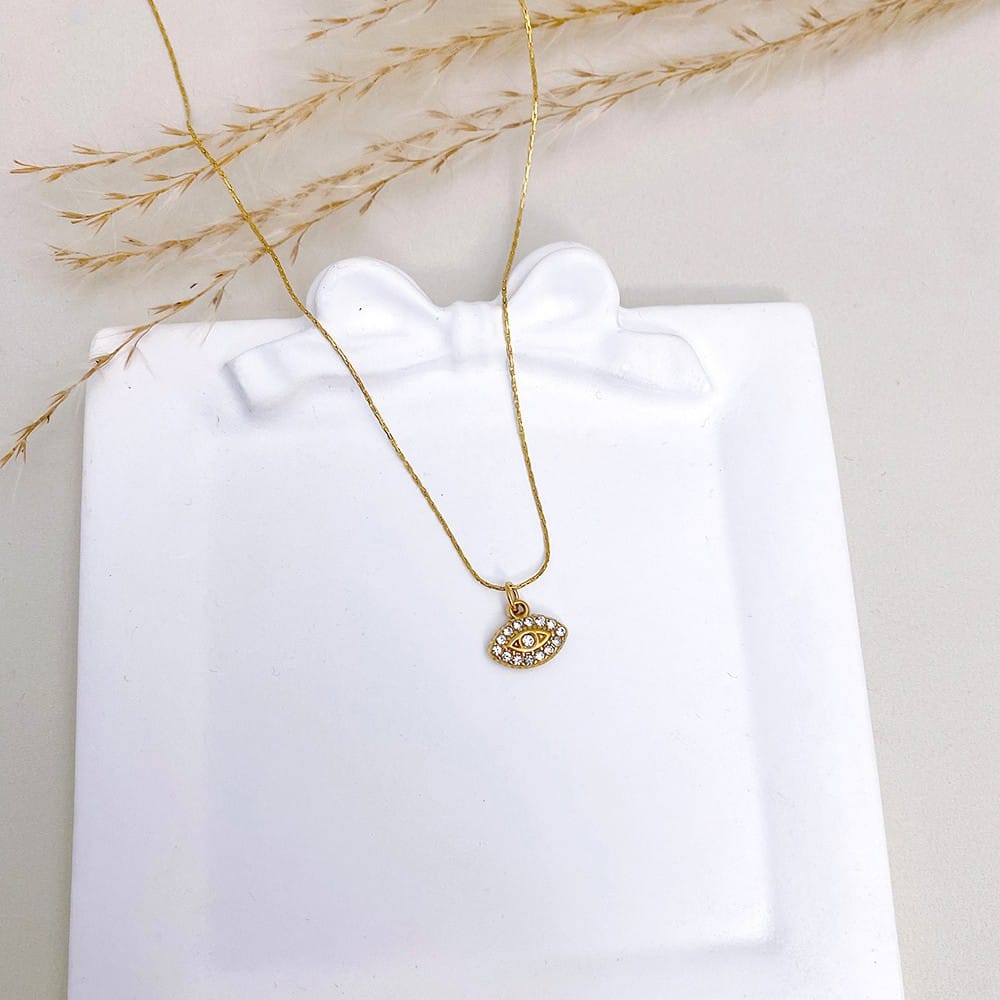 A gold chain necklace with a small evil eye pendant studded with clear rhinestones, displayed on a white gift box with dried grass stems.