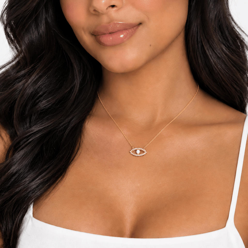 A close-up of a woman wearing a delicate gold necklace with an evil eye pendant, styled with a white spaghetti strap top against a clean white background.