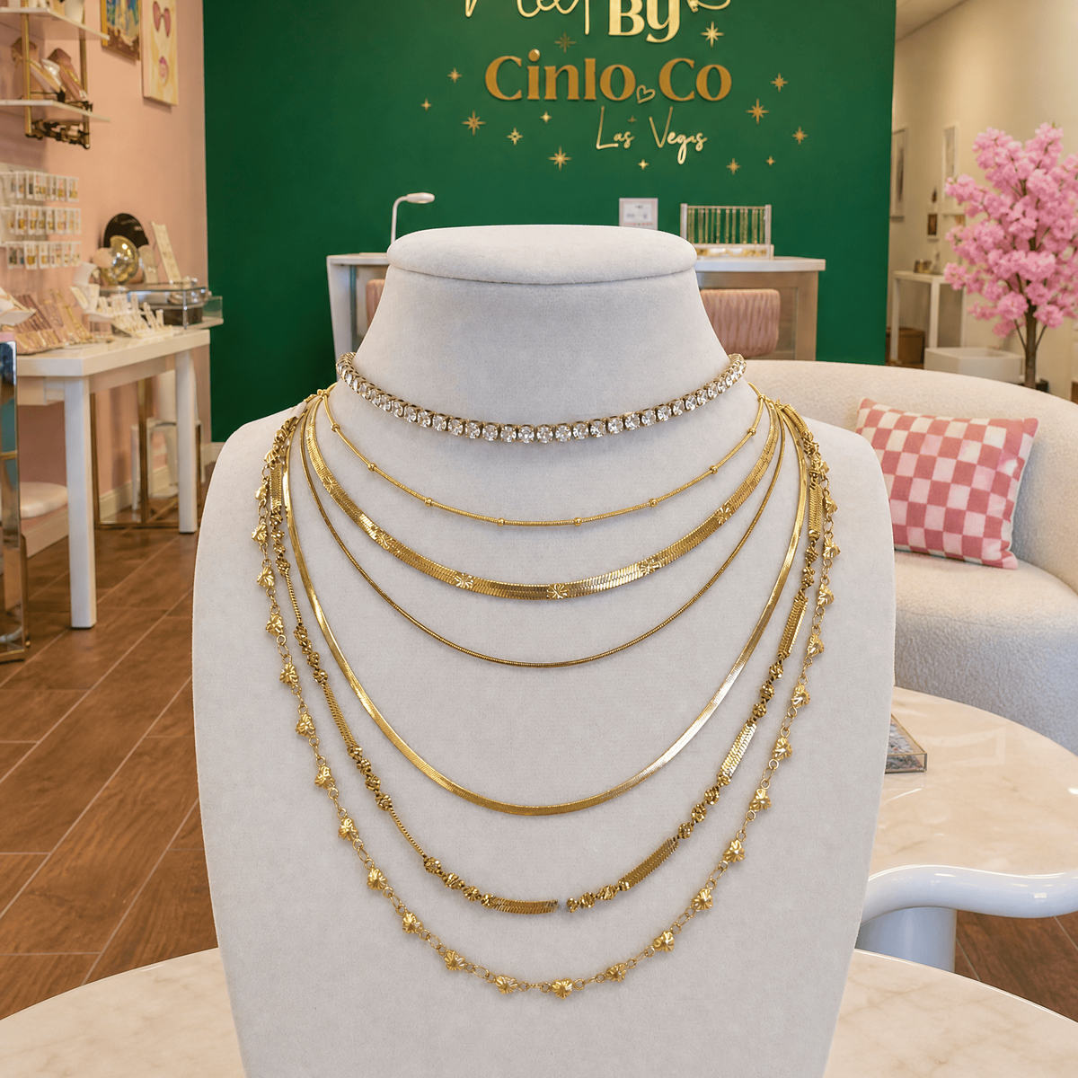 A multi-layered gold necklace set displayed on a white mannequin bust inside a jewelry store. The set includes a rhinestone choker, several gold chain layers, and a beaded gold strand.