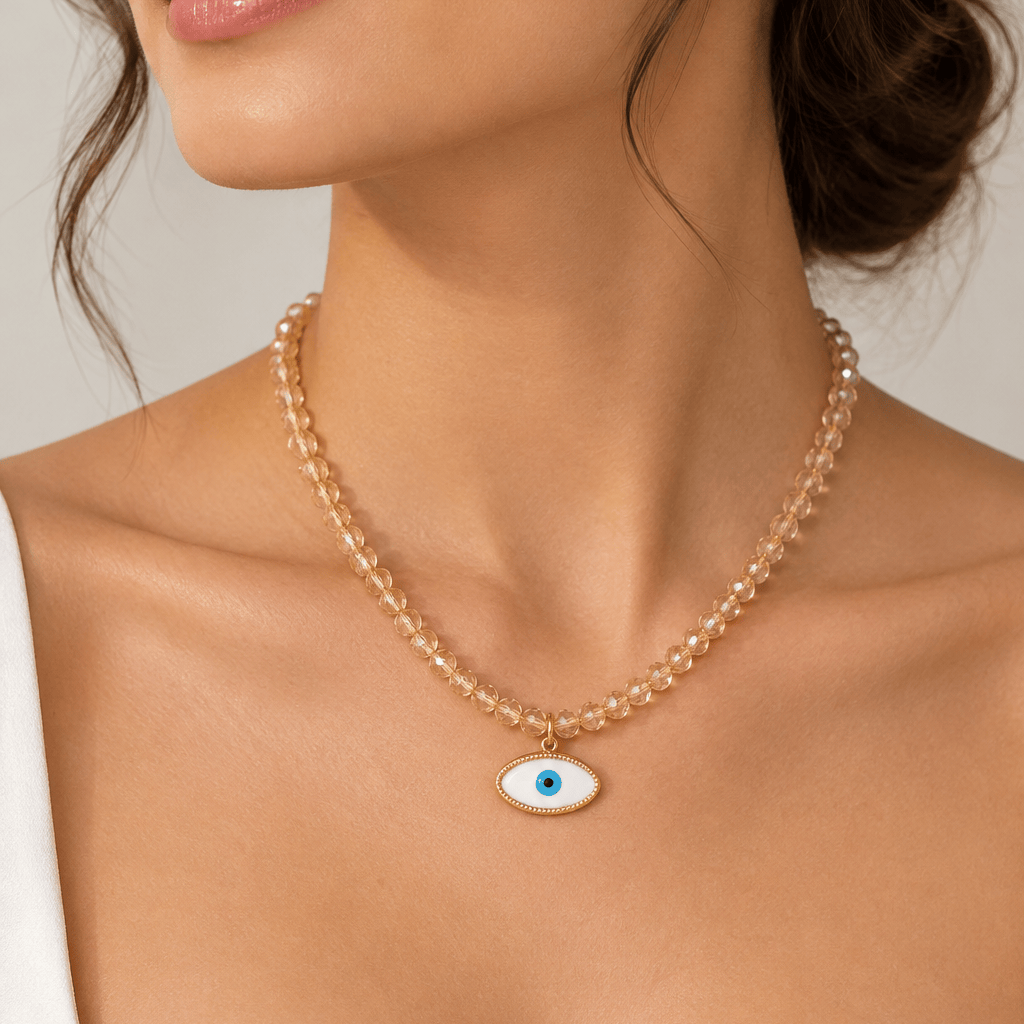 A beaded necklace with a gold-tone evil eye pendant displayed inside a pink jewelry box. The necklace features clear faceted beads and a gold-tone evil eye charm with a blue center.