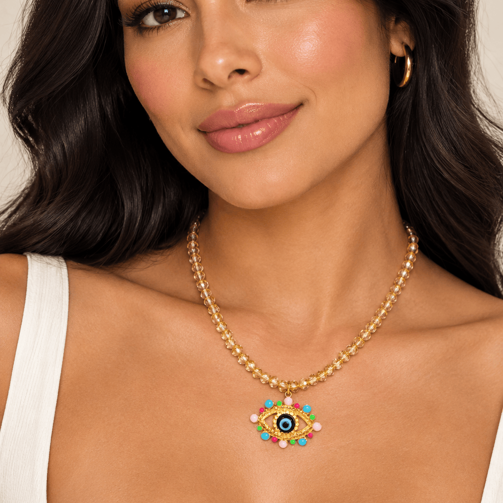 A woman wearing a gold beaded necklace with a colorful evil eye pendant, styled with a white top against a neutral beige background.
