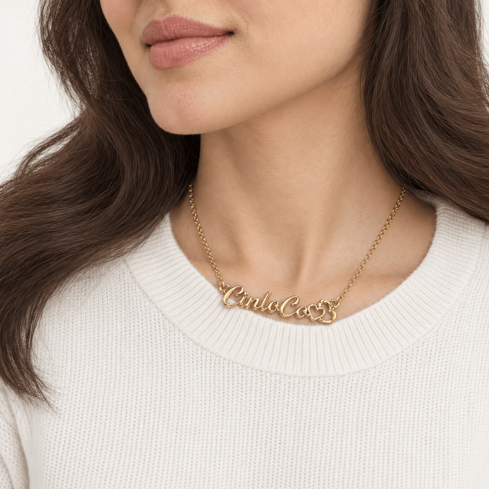 A close-up of a woman wearing a delicate gold chain necklace with a cursive nameplate reading 'CinluCo53', paired with a cream-colored ribbed knit sweater.
