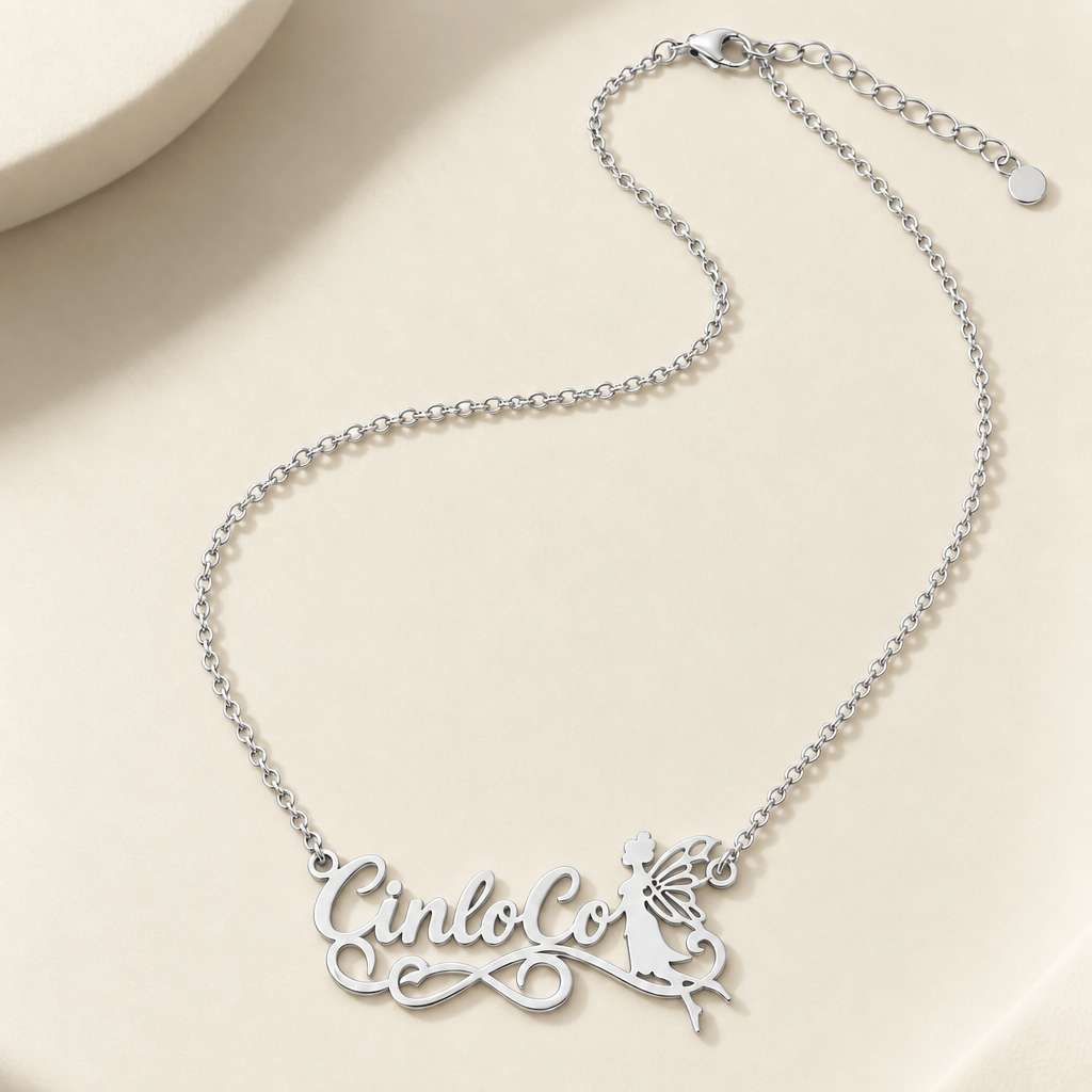 A silver nameplate necklace with a fairy cutout detail, displayed on a light beige surface with soft shadows.