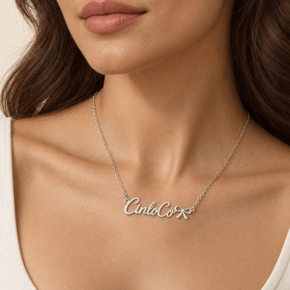 A close-up of a woman wearing a delicate silver chain necklace with a script nameplate reading 'CincoCor' in polished silver, styled with a small white flower accent.