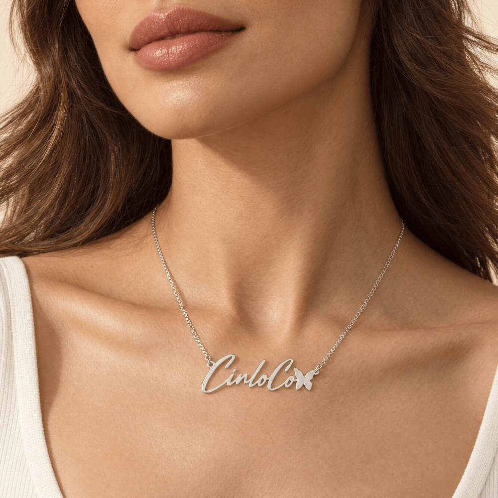 A close-up of a model wearing a delicate silver script necklace with a butterfly accent and the name 'Cinco' spelled out. The model is wearing a white ribbed tank top against a warm beige background.