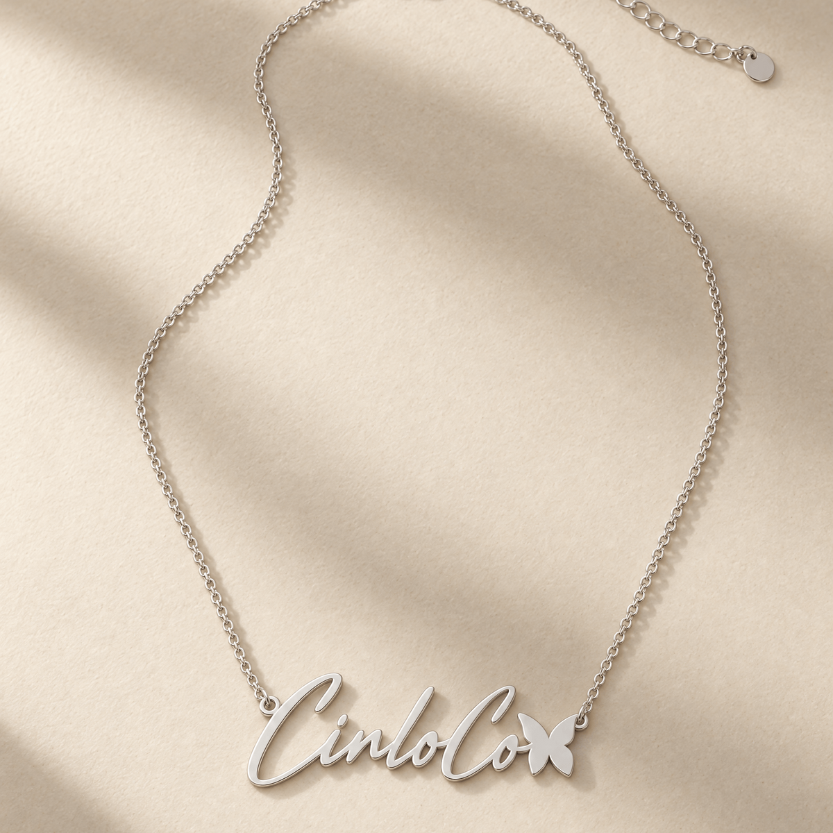A silver script name necklace reading 'Cinco' with a small butterfly charm, displayed on a textured beige background with soft diagonal shadows.