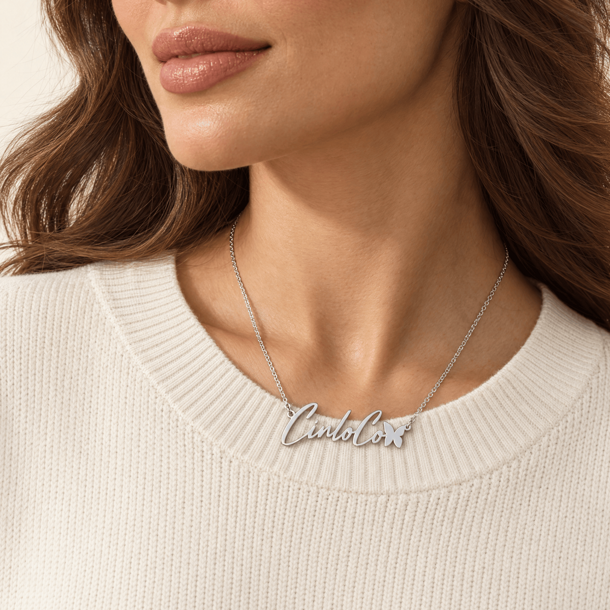 A close-up of a woman wearing a silver script name necklace that reads 'Candela' with a small flower accent, paired with a cream ribbed knit sweater.