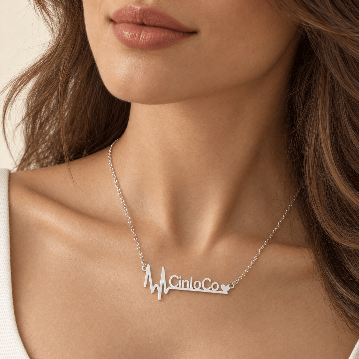 A close-up of a woman wearing a silver necklace with a heartbeat line design and the text 'Cinco de Co' with a small heart at the end, paired with a white scoop-neck top.