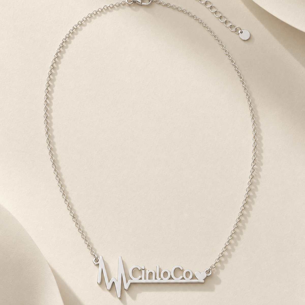 A silver chain necklace with a custom nameplate spelling 'Cinco Co' in a heartbeat line design, displayed on a beige fabric background.
