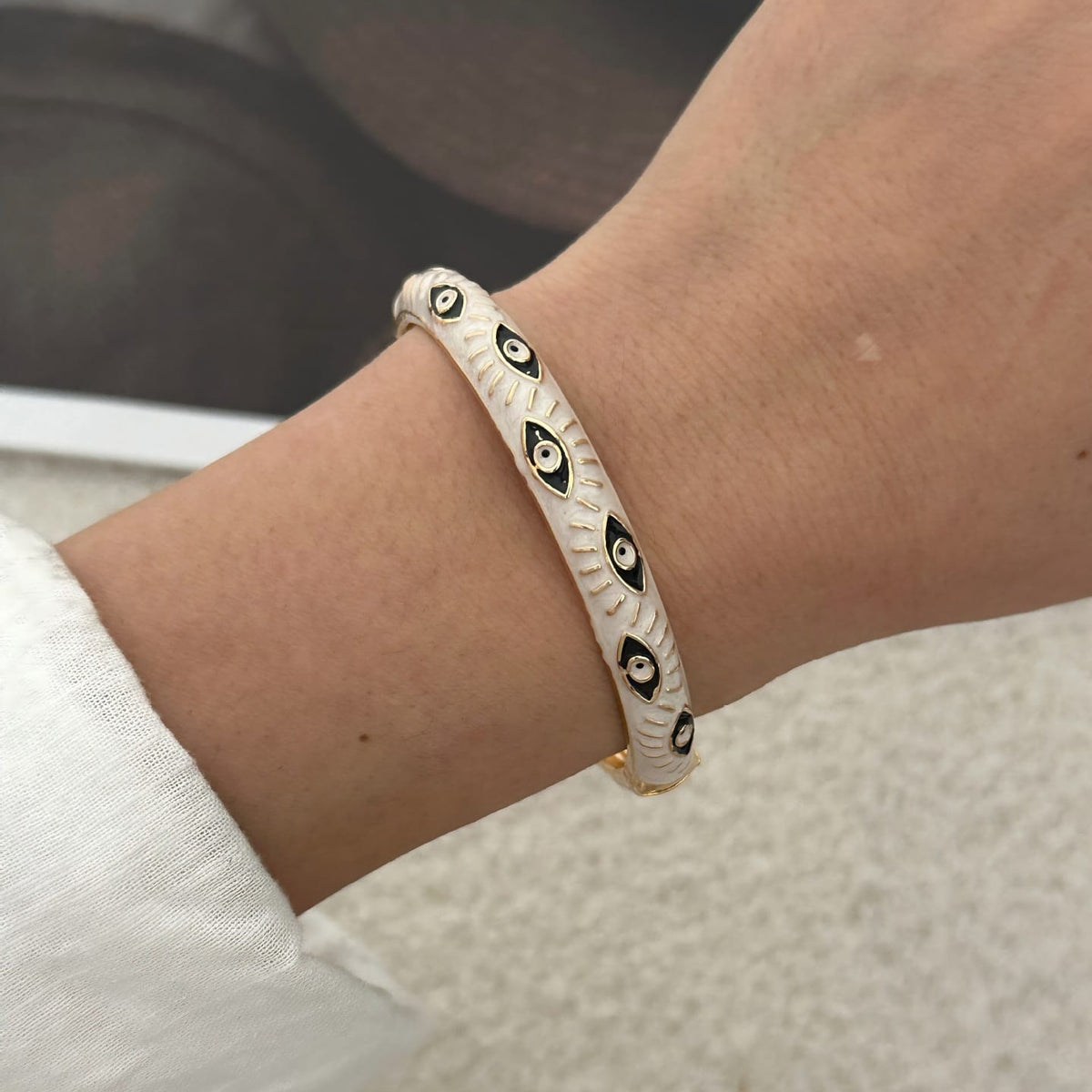 A close-up of a person's wrist wearing a white beaded bracelet with black and white eye motifs and a small gold clasp, set against a soft beige textured background.