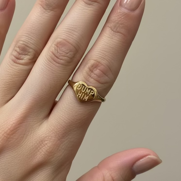 Hand wearing a gold ring with 'DUMP HIM' on a neutral background