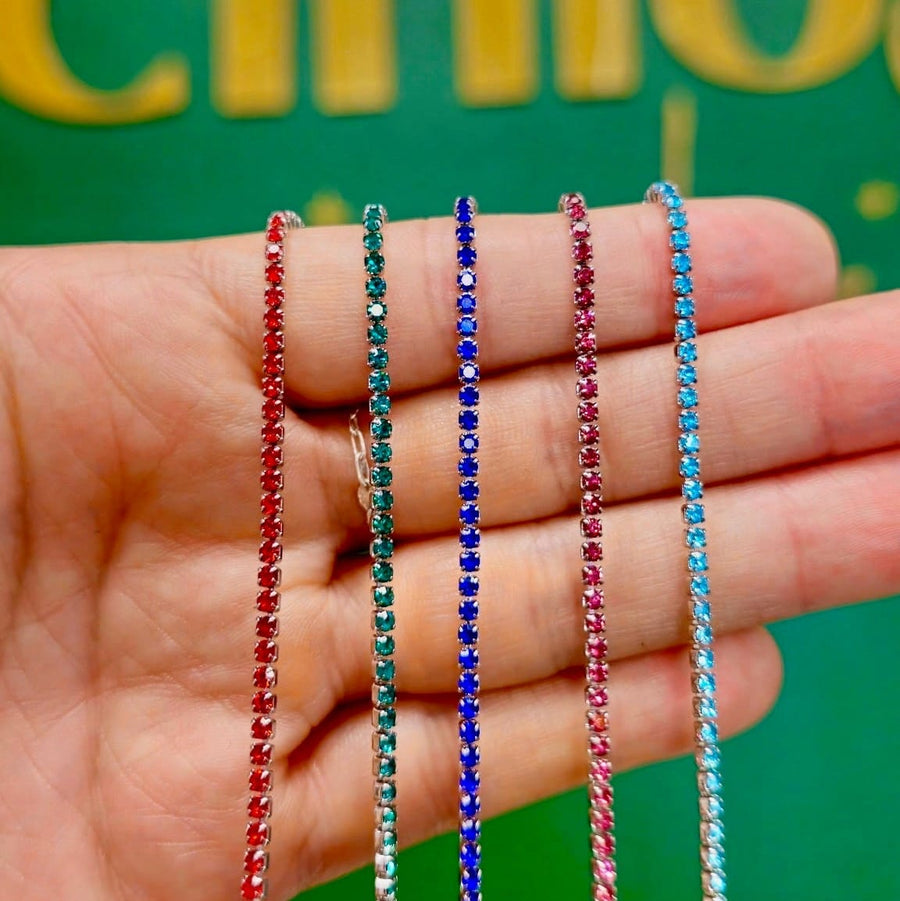 Five thin beaded necklaces in red, teal, dark blue, pink, and light blue are draped across an open hand, with a green sign featuring gold lettering blurred in the background.