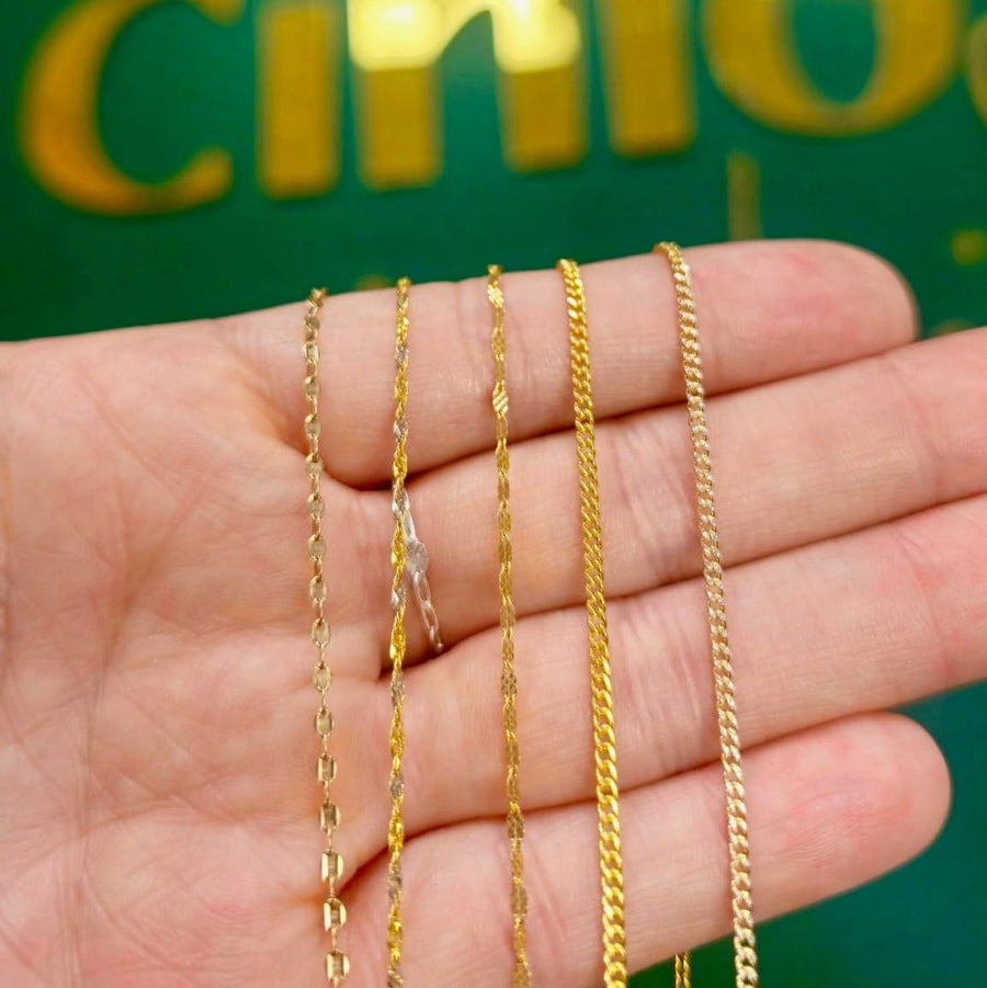 Four gold-tone chain necklaces of varying link styles held in a person's hand, with a green background featuring gold lettering.