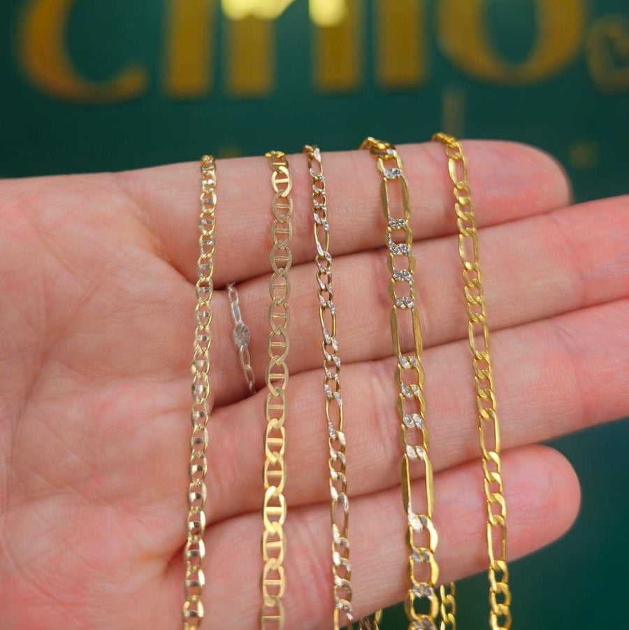 Four gold-toned chains of varying link styles held in a person's hand, with a dark green and gold blurred background featuring the text 'Mott By Cinco'.