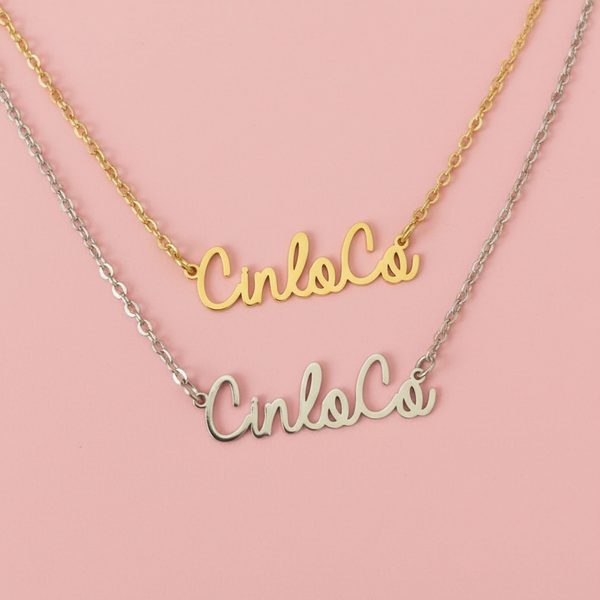 Two nameplate necklaces, one gold and one silver, on a pink background.