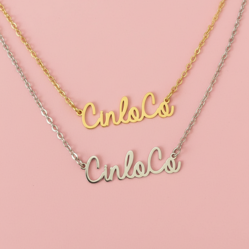 Two nameplate necklaces, one gold and one silver, on a pink background.