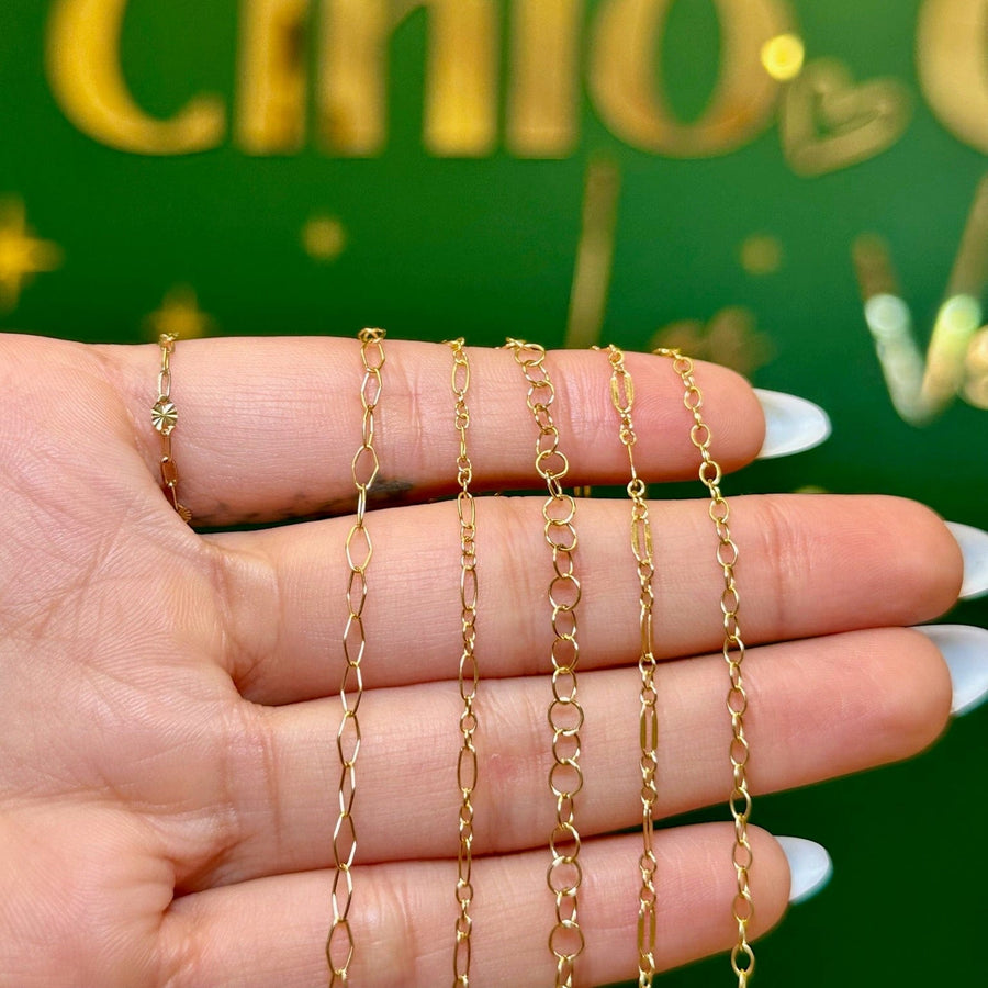 A hand with white manicured nails holds four delicate gold chains of varying link styles against a green background with gold lettering.