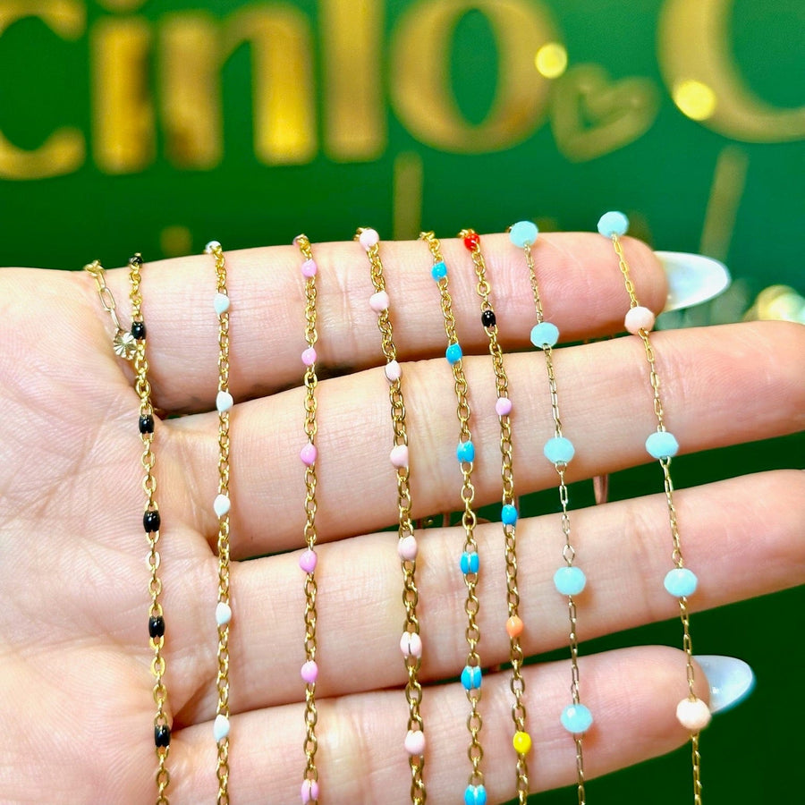 A hand holding six delicate gold chain necklaces with small colorful beads, set against a green background with gold lettering.