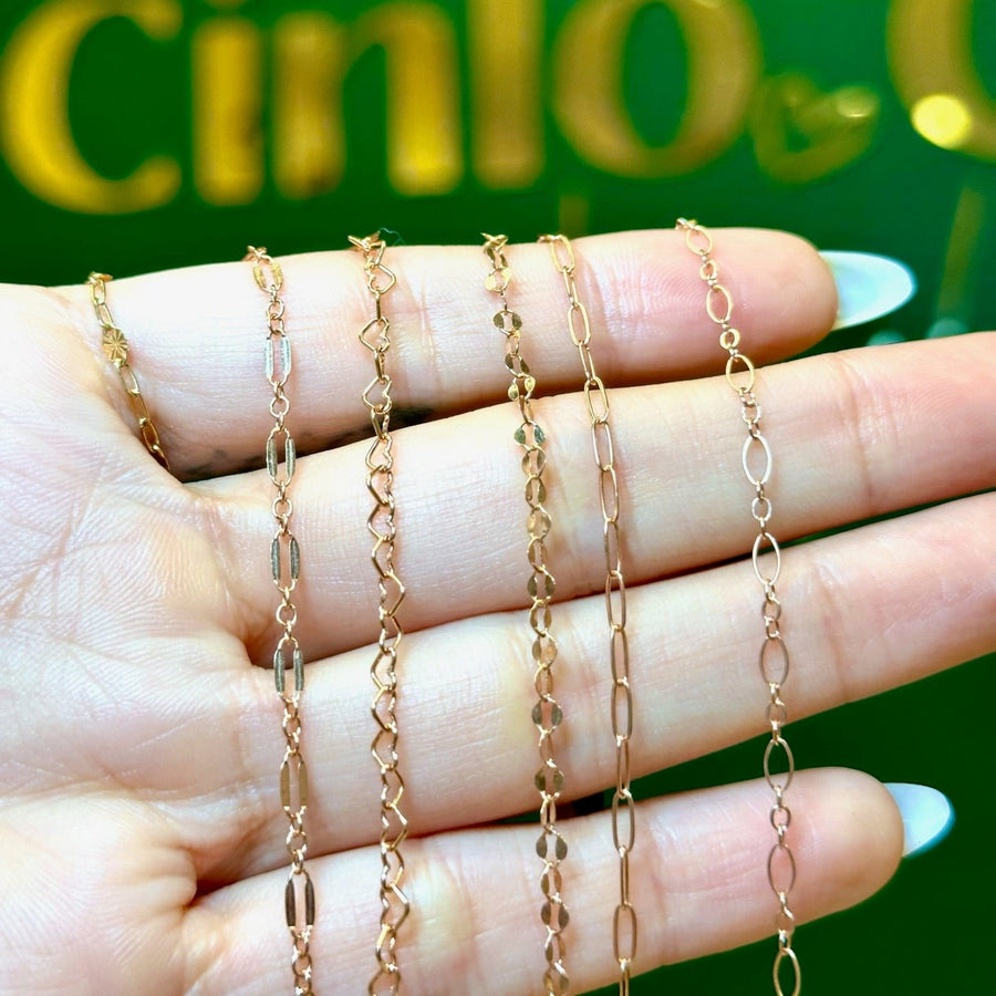 Four delicate rose gold chains of different link styles draped over a hand with white manicured nails, set against a green background with gold lettering.
