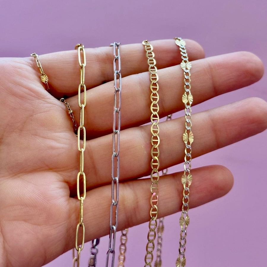 Four delicate chains in gold and silver tones draped over an open hand against a solid purple background.