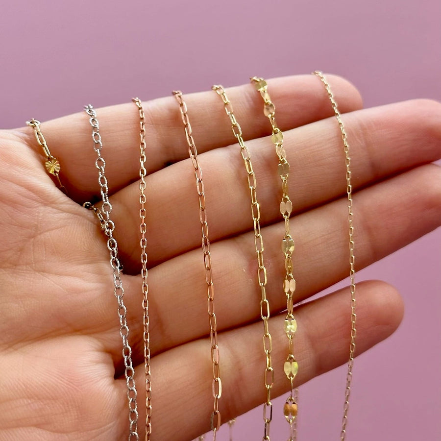 Four delicate chain necklaces in gold and silver tones draped over a person's hand against a solid pink background.