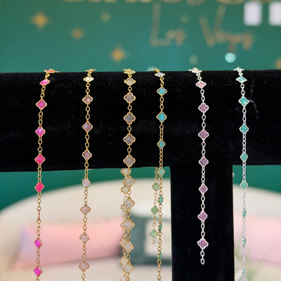 Five delicate chain bracelets with alternating pink and clear gemstone-like beads are displayed on a black velvet jewelry stand. The background features a green wall with gold lettering that reads 'By Cinio Co.