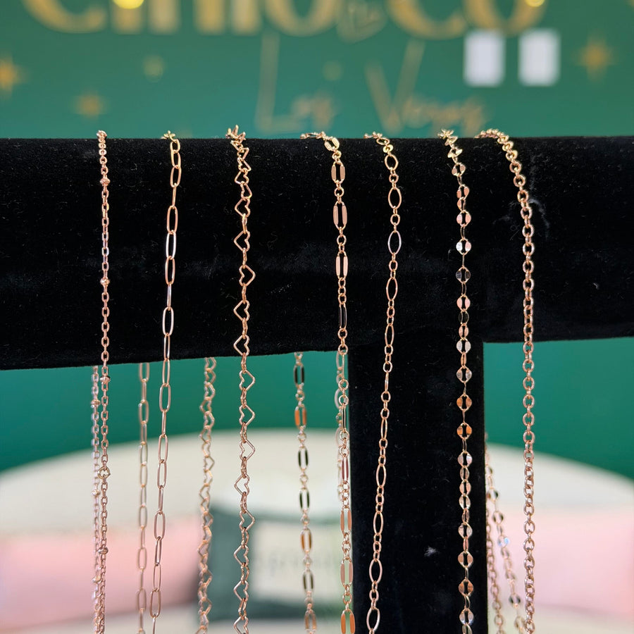 Six rose gold chain necklaces in different link styles are displayed hanging from a black velvet jewelry stand. The background features a green sign with gold lettering that reads 'By Cinco Co'.