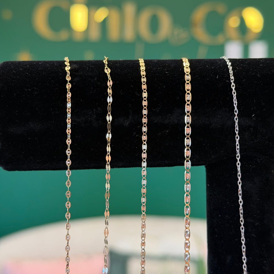 Four delicate chain necklaces in gold, rose gold, and silver tones displayed on a black velvet jewelry stand, with a green and gold blurred background featuring the text 'Mia by Cinla Co'.