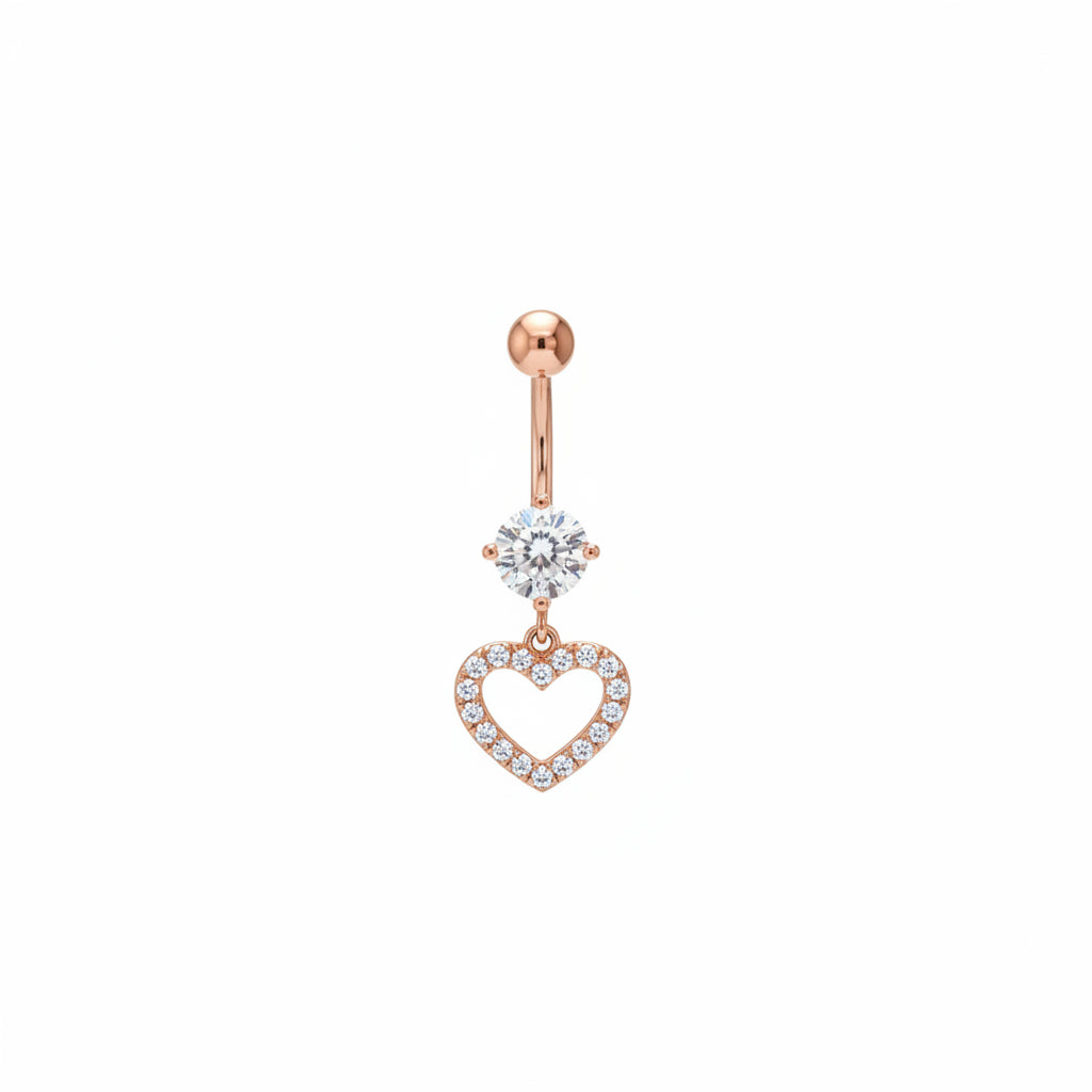 A hand with manicured nails holds a rose gold hoop earring featuring a round clear crystal and a dangling heart-shaped crystal charm, set against a blurred boutique background.