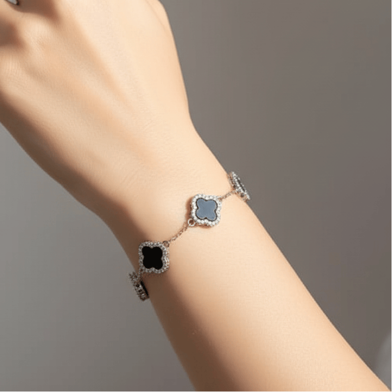 A silver-toned chain bracelet with three clover-shaped charms, worn on a model's wrist. The top and bottom of each clover are outlined with small clear stones, and the center of each charm is a dark blue-gray color.