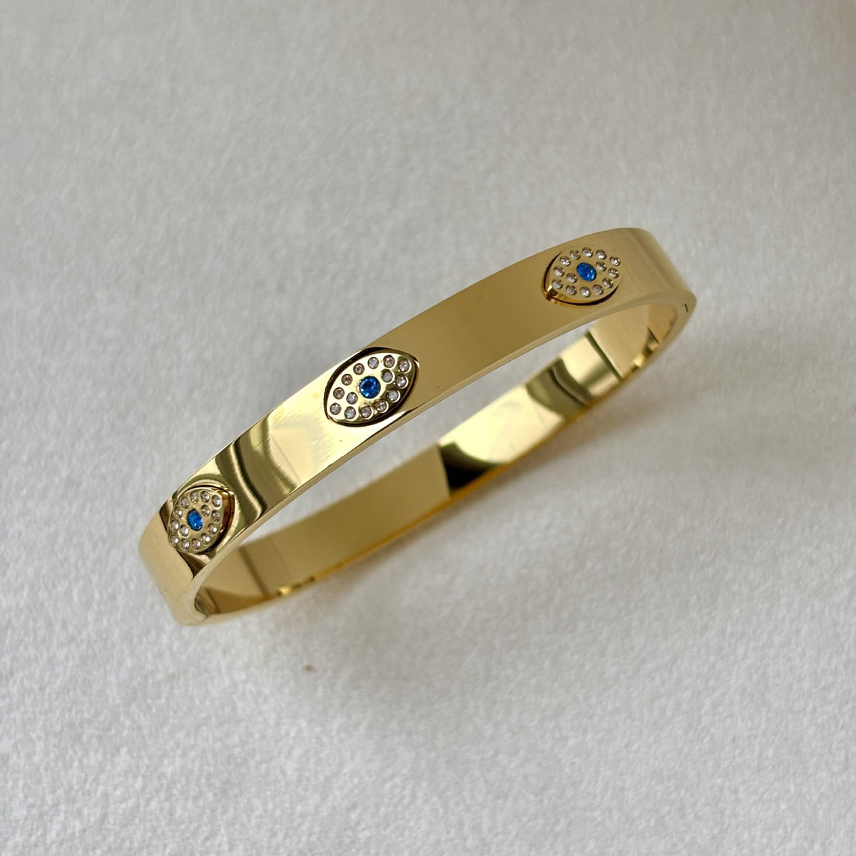 A polished gold bangle bracelet featuring three evil eye motifs set with small white stones and blue accents, displayed on a textured white background.