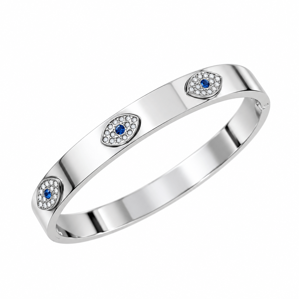 A polished silver-toned metal bangle bracelet featuring three oval evil eye motifs with small blue stones and clear crystals, displayed on a textured cream background.