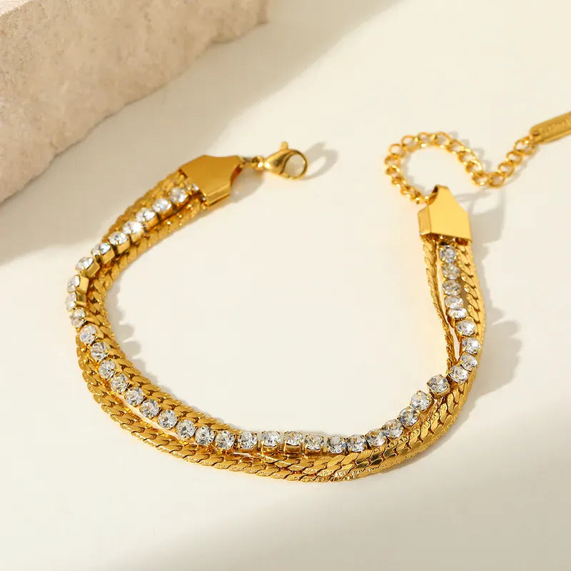 A gold-toned chain bracelet with multiple rows of clear rhinestones, displayed on a light cream surface with a soft beige textured prop in the corner.