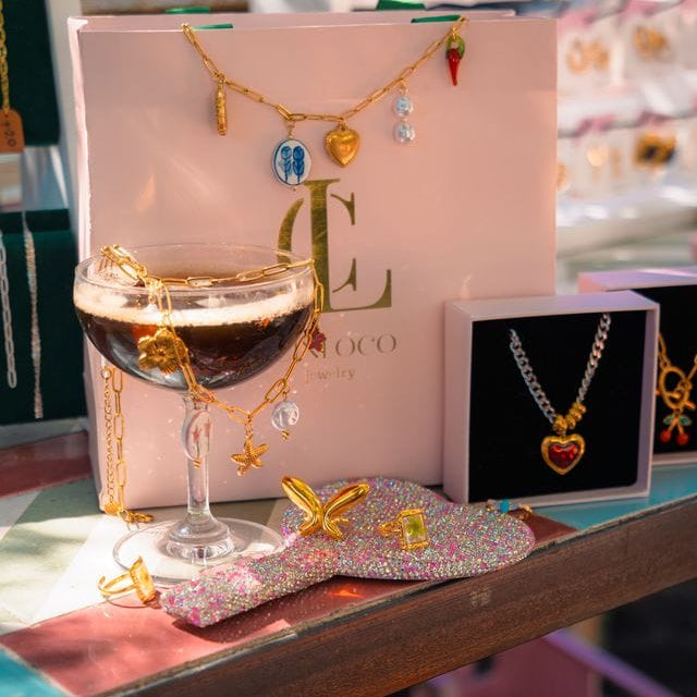 Jewelry set with necklaces and a glass of red wine on a pink box with 'L' logo.