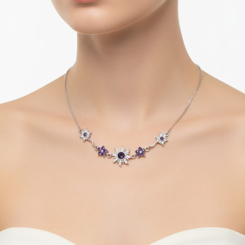 A close-up of a model wearing a delicate silver necklace with a series of flower-like settings, some holding clear stones and others holding deep purple stones, paired with a white strapless top against a clean white background.