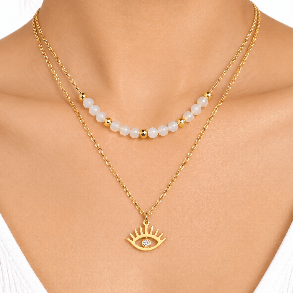 A close-up of a model's neck and chest wearing a double-layered gold necklace. The top layer features a delicate chain with small white pearl-like beads separated by tiny gold spacer beads.