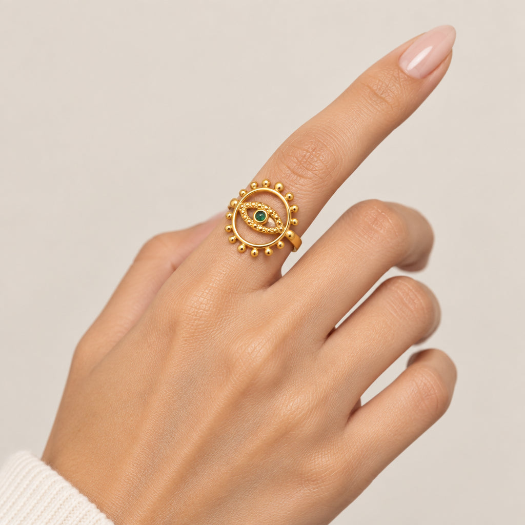 A gold-toned ring with an evil eye motif and a small green stone, worn on a person's finger against a soft beige background.