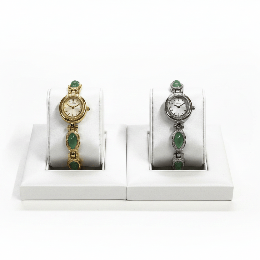 Two vintage-style wristwatches displayed on white stand-in forms against a white background. The watch on the left is gold-toned with green stones, and the watch on the right is silver-toned with green stones.