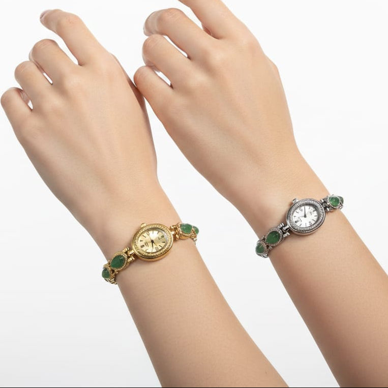Two women's watch bracelets shown on a model's wrists, one in yellow gold and one in silver, both featuring green stone accents on a white background.