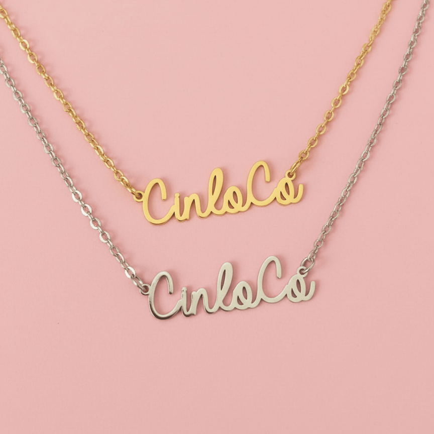 Two nameplate necklaces, one gold and one silver, on a pink background.