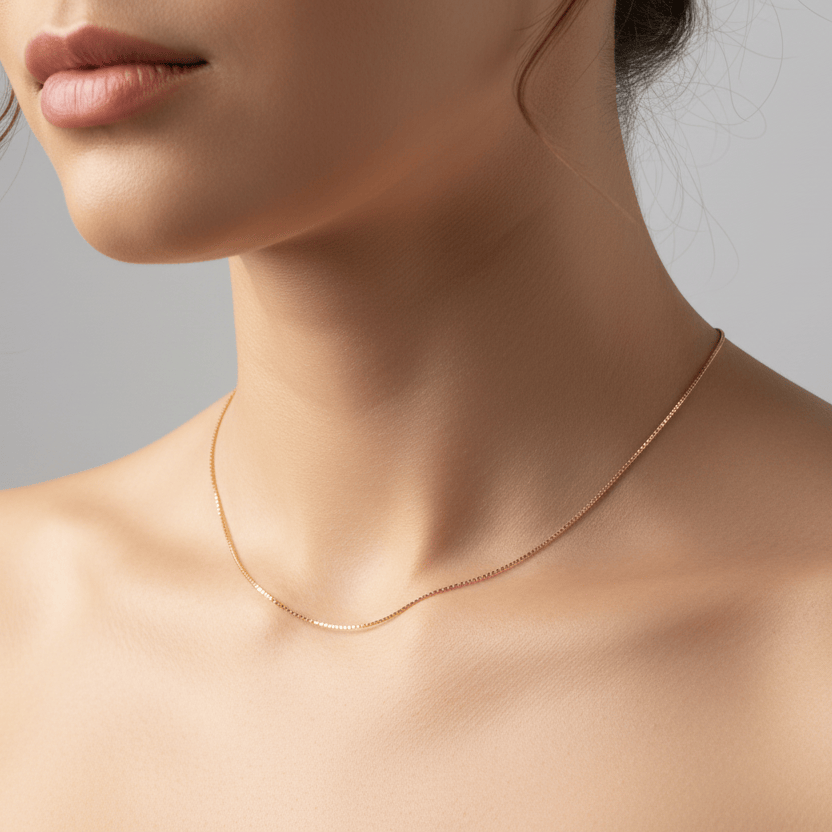 A close-up of a model's neck and upper chest wearing a delicate rose gold chain necklace against a soft gray background.
