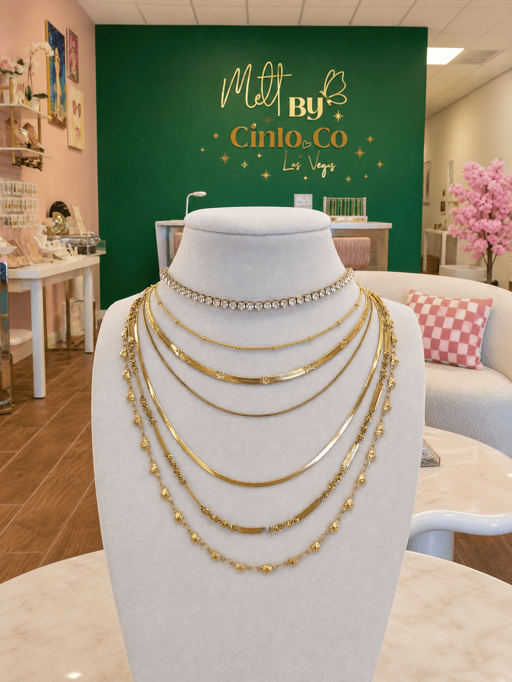 A multi-layered gold necklace set displayed on a white mannequin bust inside a jewelry store. The set includes a rhinestone choker, several gold chain layers, and a beaded gold strand.