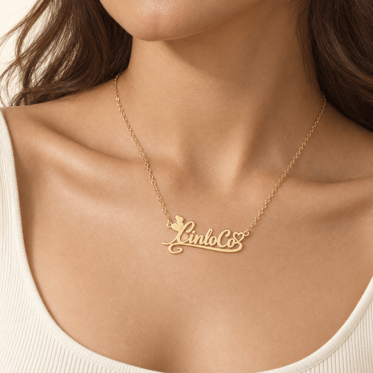 A close-up of a model wearing a delicate gold chain necklace with a personalized script pendant reading 'Lindo Co' and a small butterfly accent, styled with a cream ribbed scoop-neck top.