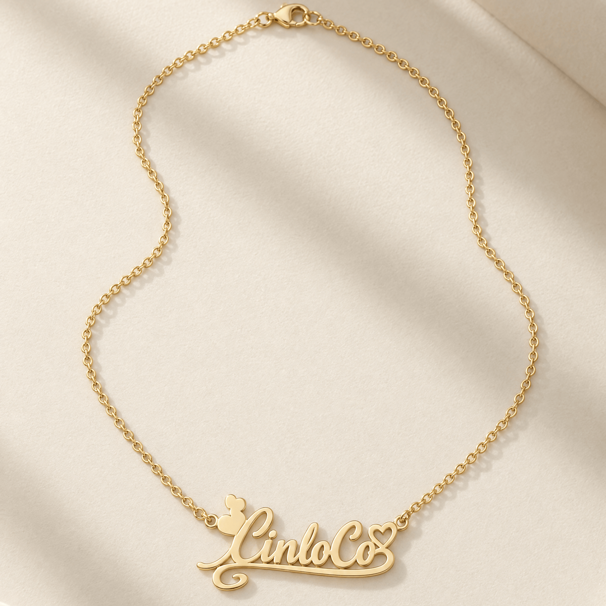 A gold chain necklace with a cursive nameplate reading 'Lindo Co' and a small butterfly motif, displayed on a soft cream satin fabric background.